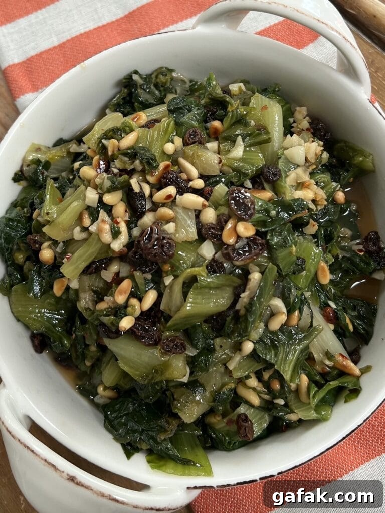 Sautéed Italian Escarole with pine nuts and raisins in white dish over decorative towel.