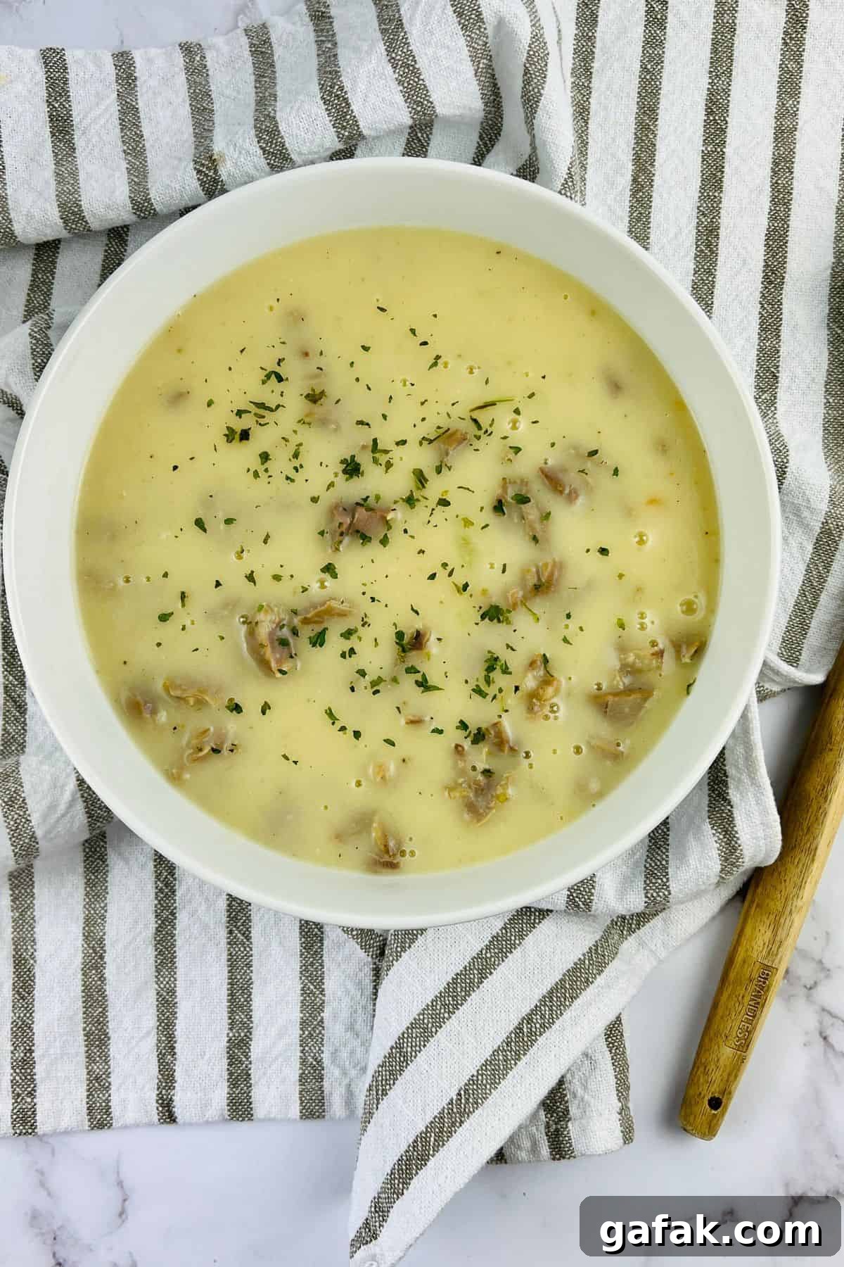 A white bowl of creamy sauce garnished with herbs, placed on a striped towel with a wooden-handled spoon beside it.
