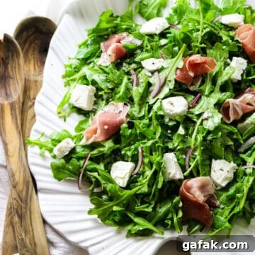 rocket salad with arugula leaves on a white platter with goat cheese and prosciutto toppings with wooden spoons on the left.