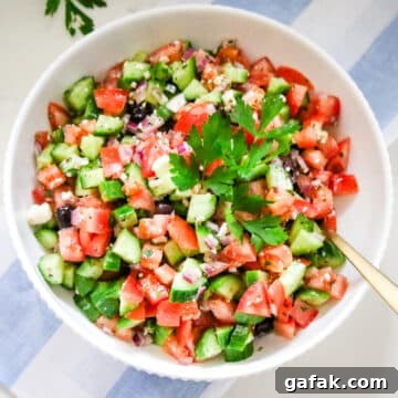 large bowl of Greek salad being tossed with a herb vinaigrette, with the dressing visible coating the cucumbers and tomatoes.