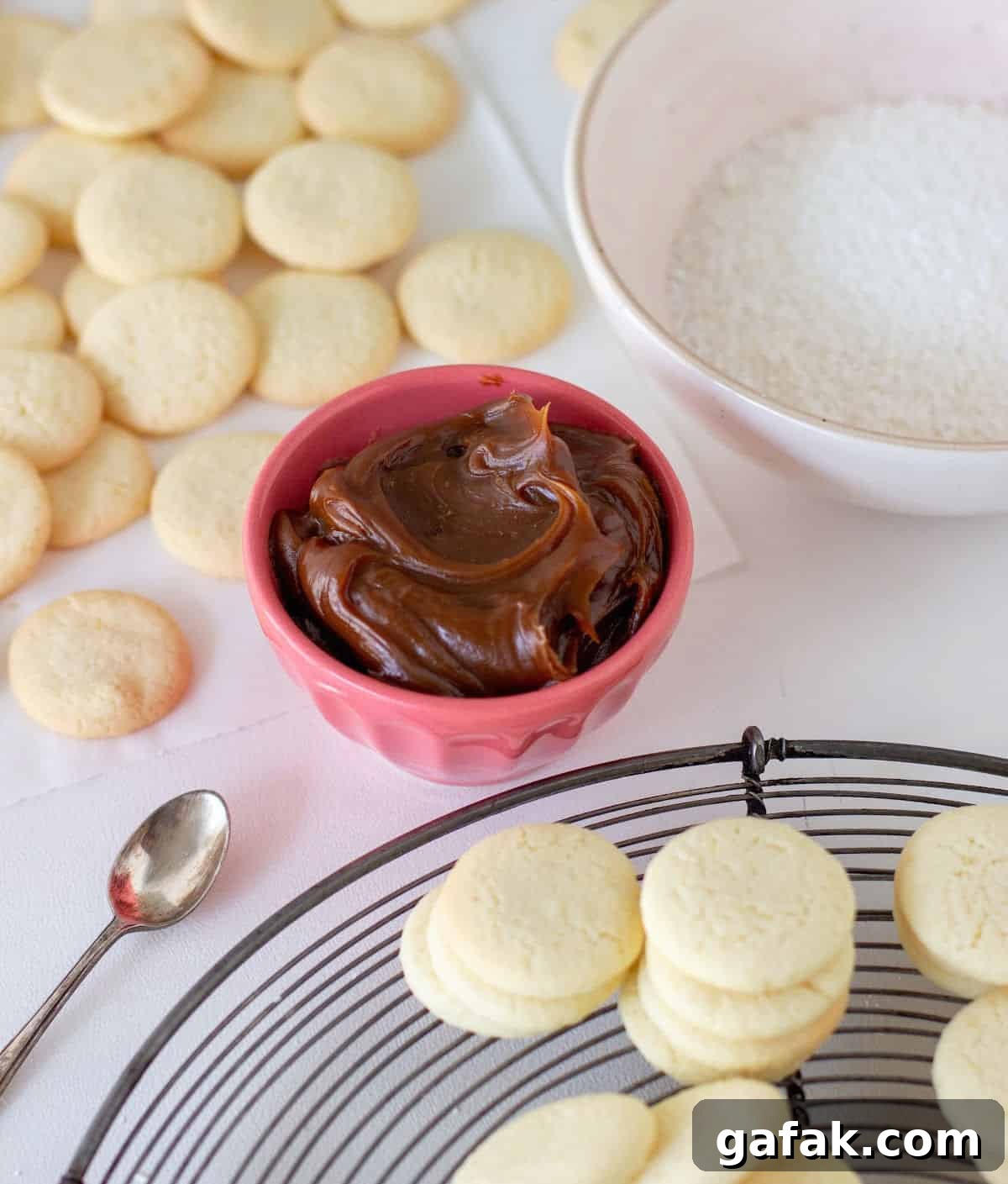 Cornstarch cookies on wire rack, dulce de leche in pink bowl, coconut in bowl, white surface.