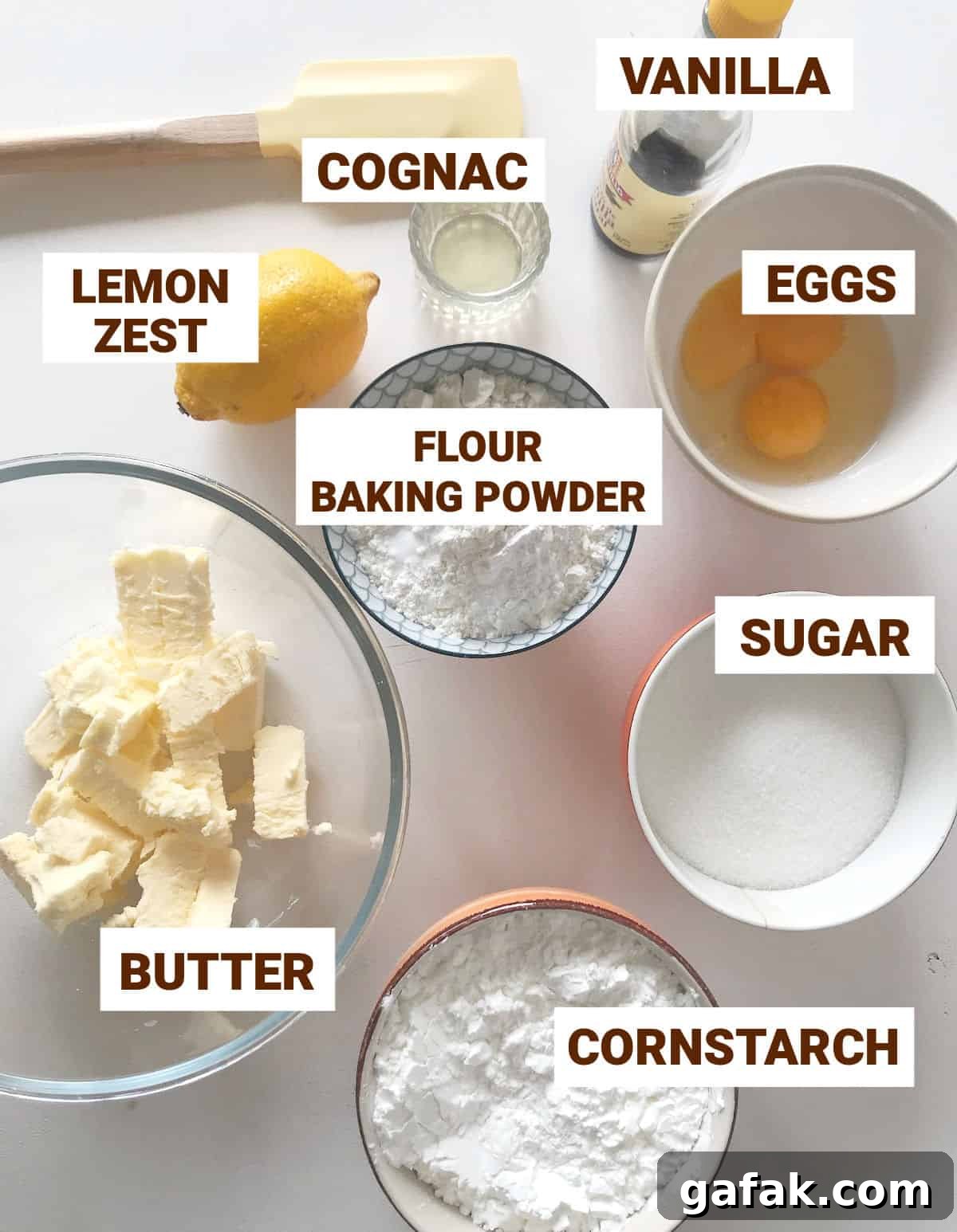 Ingredients for cornstarch alfajores in bowls on white surface, yellow spatula, whole lemon, butter, eggs, sugar, vanilla