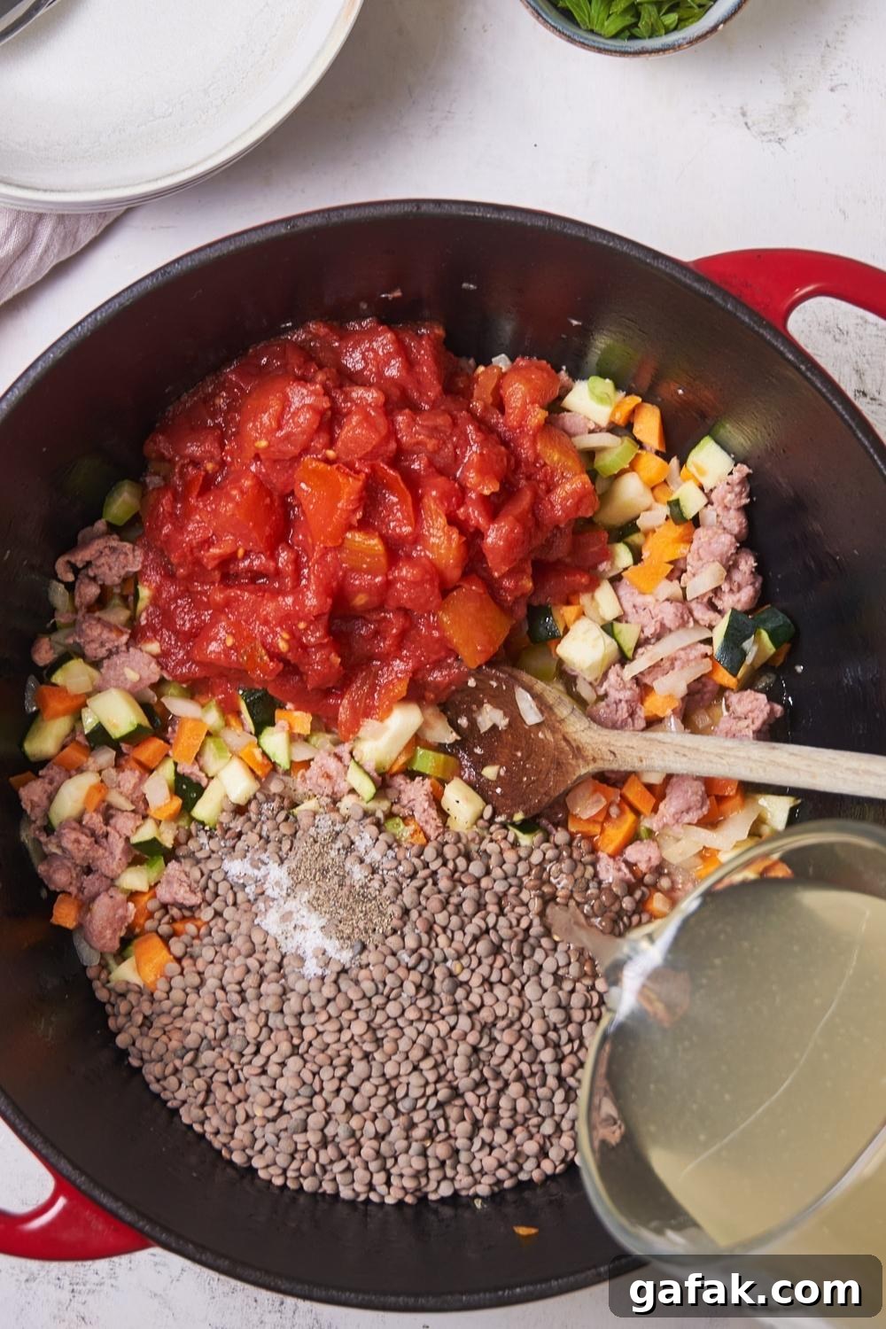 A large pot with veggies, sausage, diced tomatoes, lentils, and a measuring cup pouring chicken broth into the pot.