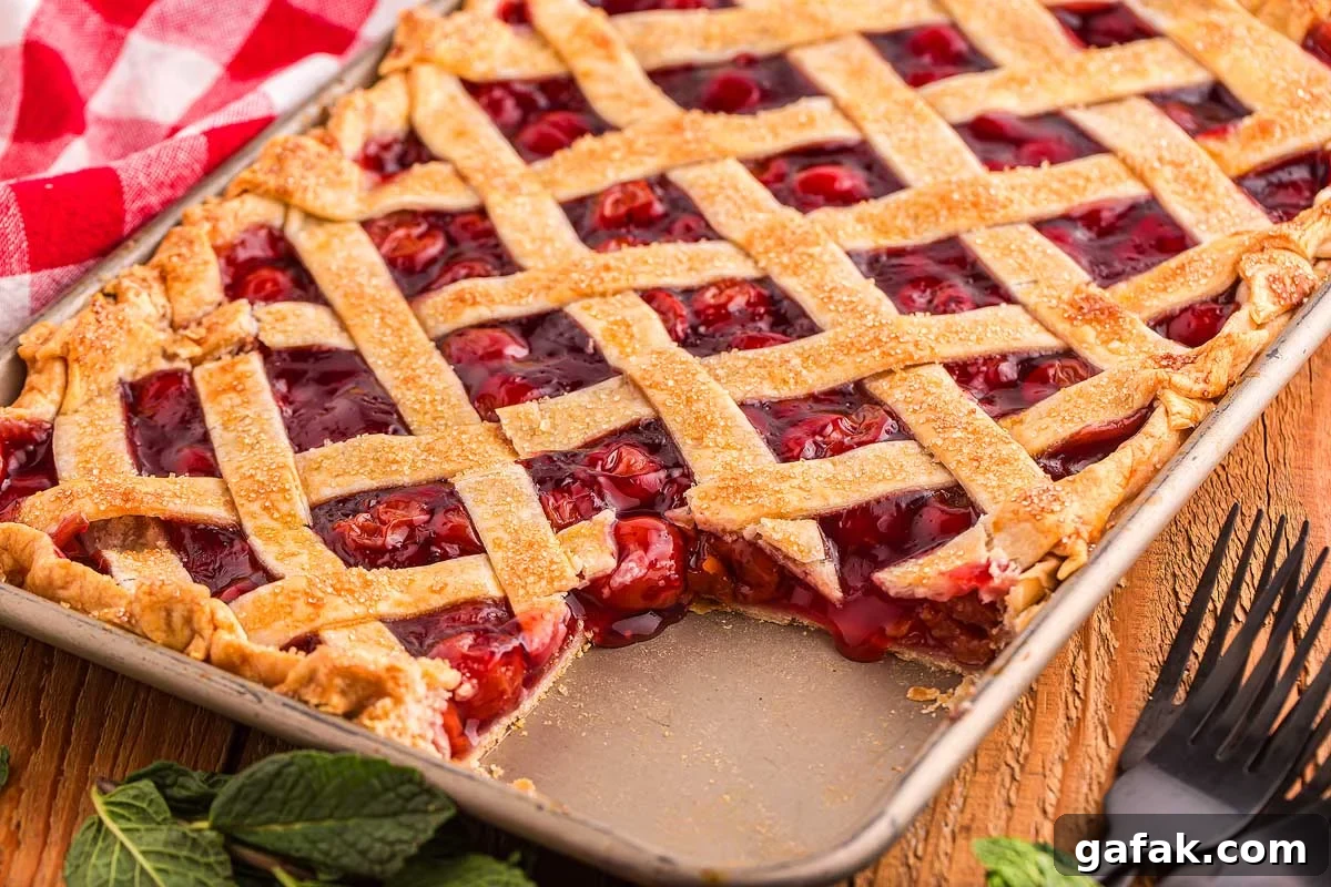 Cherry slab pie with a lattice crust rests in a baking pan, one slice missing. A red checkered cloth lies nearby, adding a touch of rustic charm.
