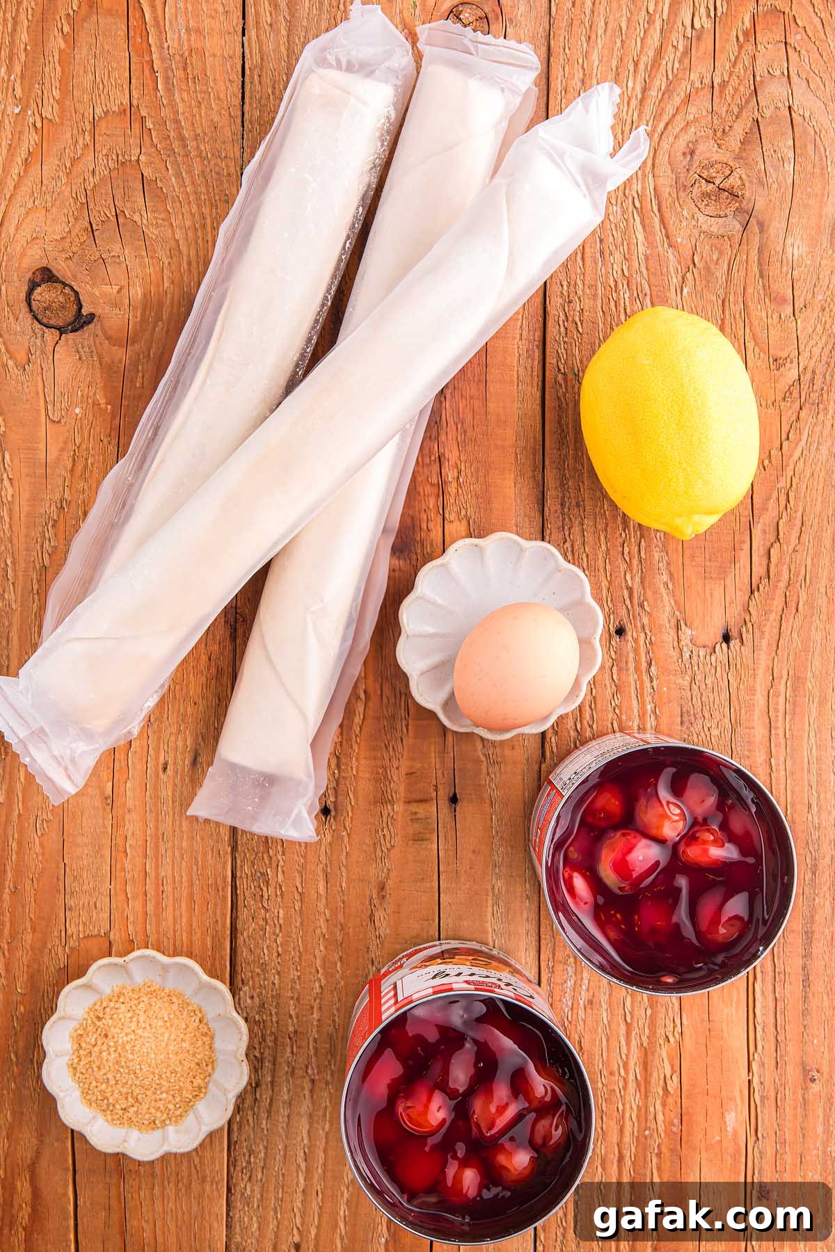 Top view of phyllo dough, a lemon, an egg, sugar, and two glasses of red punch with ice on a wooden surface—perfect ingredients to start crafting a cherry slab pie masterpiece.
