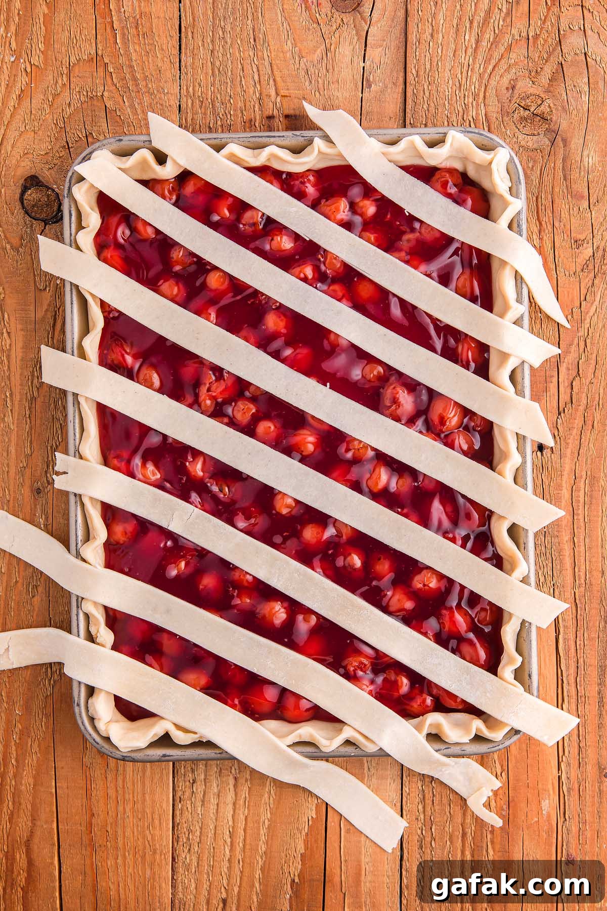Unbaked cherry slab pie with lattice strips rests on a wooden surface.