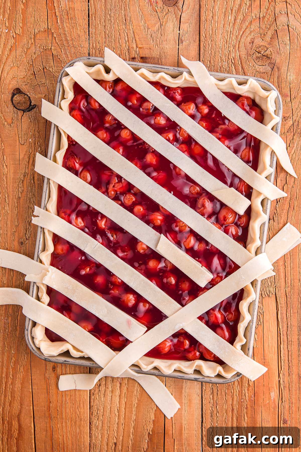 Cherry slab pie with a lattice of uncooked crust strips rests on a wooden surface, ready for baking.