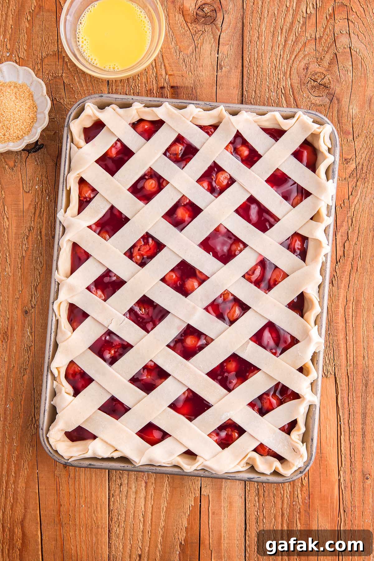 A delightful cherry slab pie with a lattice crust sits on a wooden table, accompanied by a bowl of egg wash and sugar.