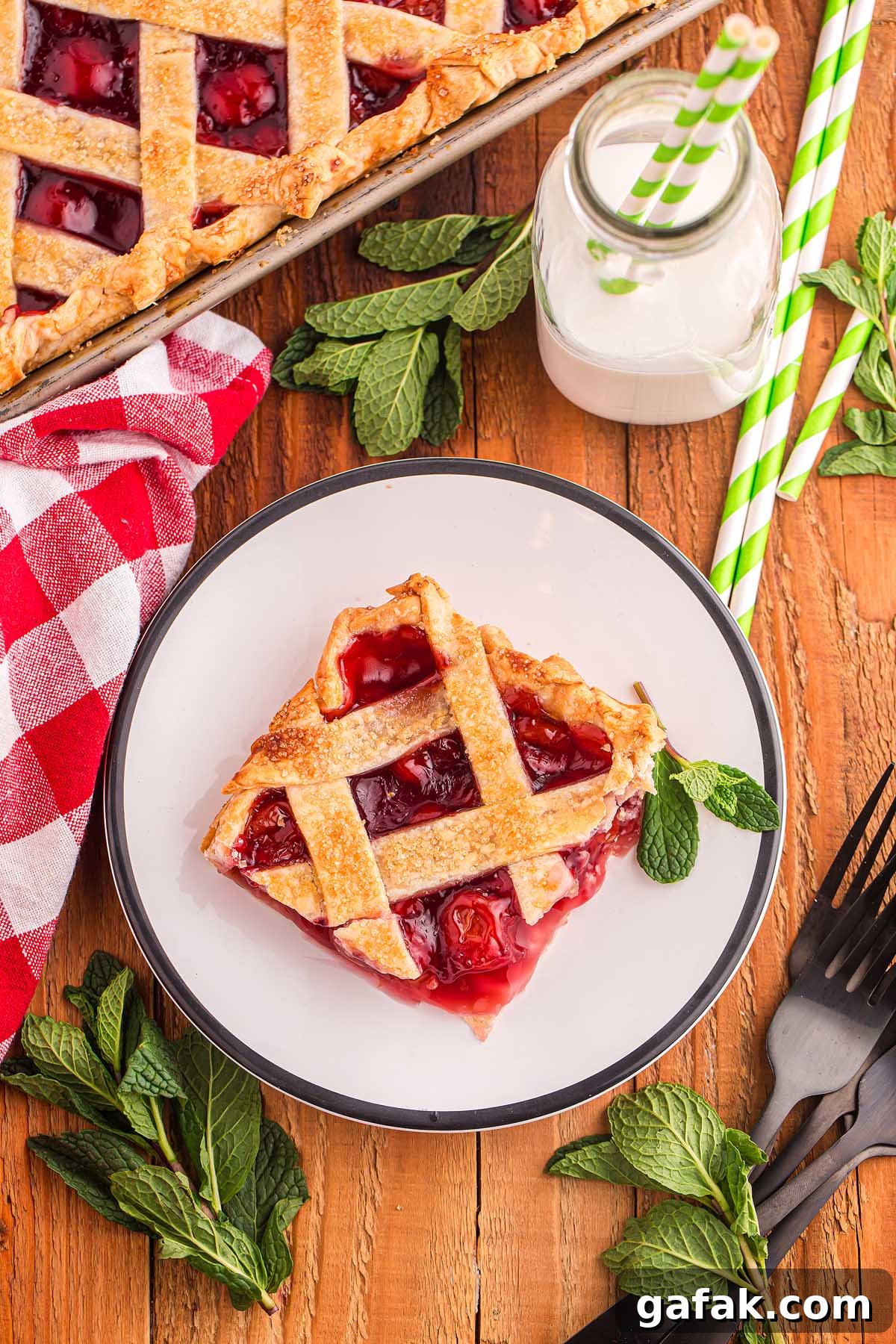A slice of cherry slab pie with a lattice crust sits invitingly on a plate, surrounded by fresh mint leaves and set against a checkered napkin. Nearby, an old-fashioned milk bottle completes the charming tableau.