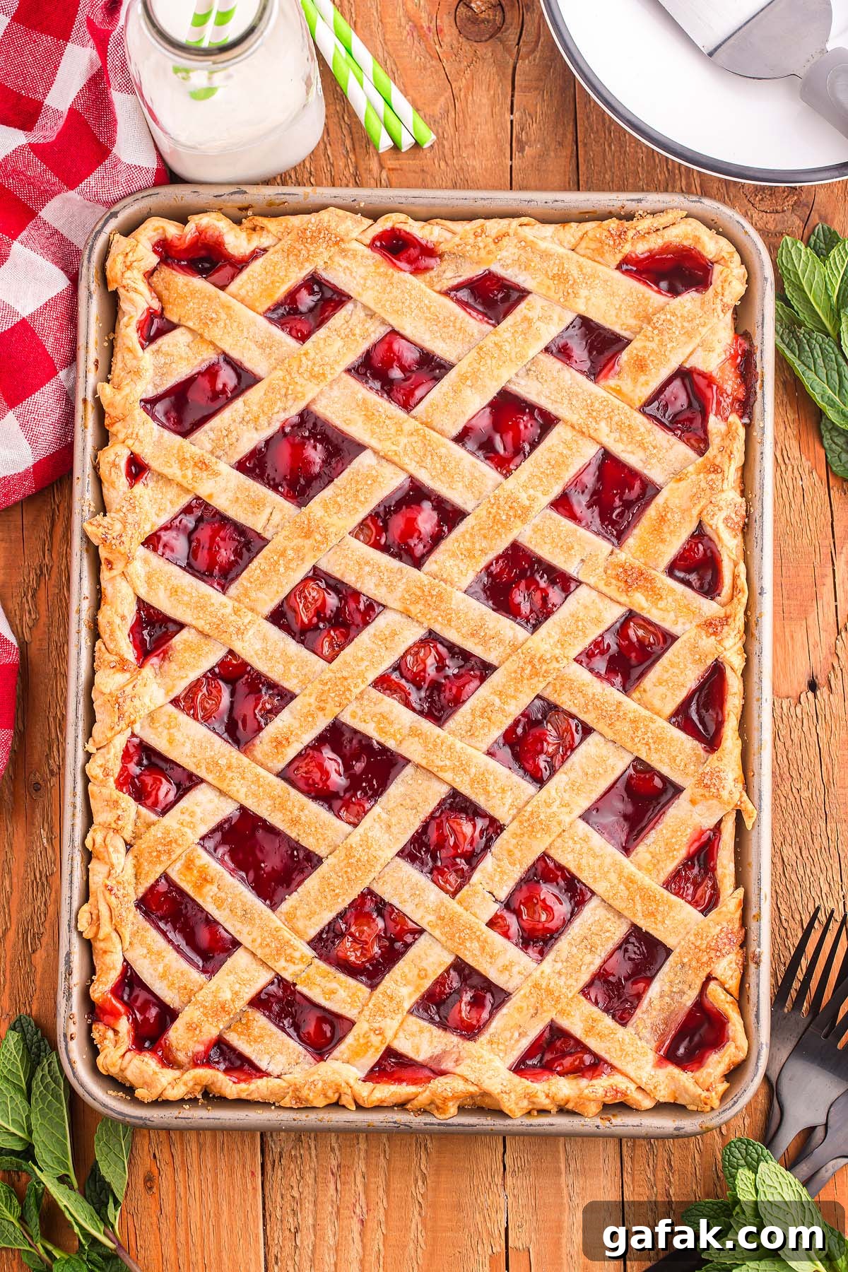 A cherry slab pie with a lattice crust rests on a wooden table, surrounded by mint leaves and a mason jar of milk.
