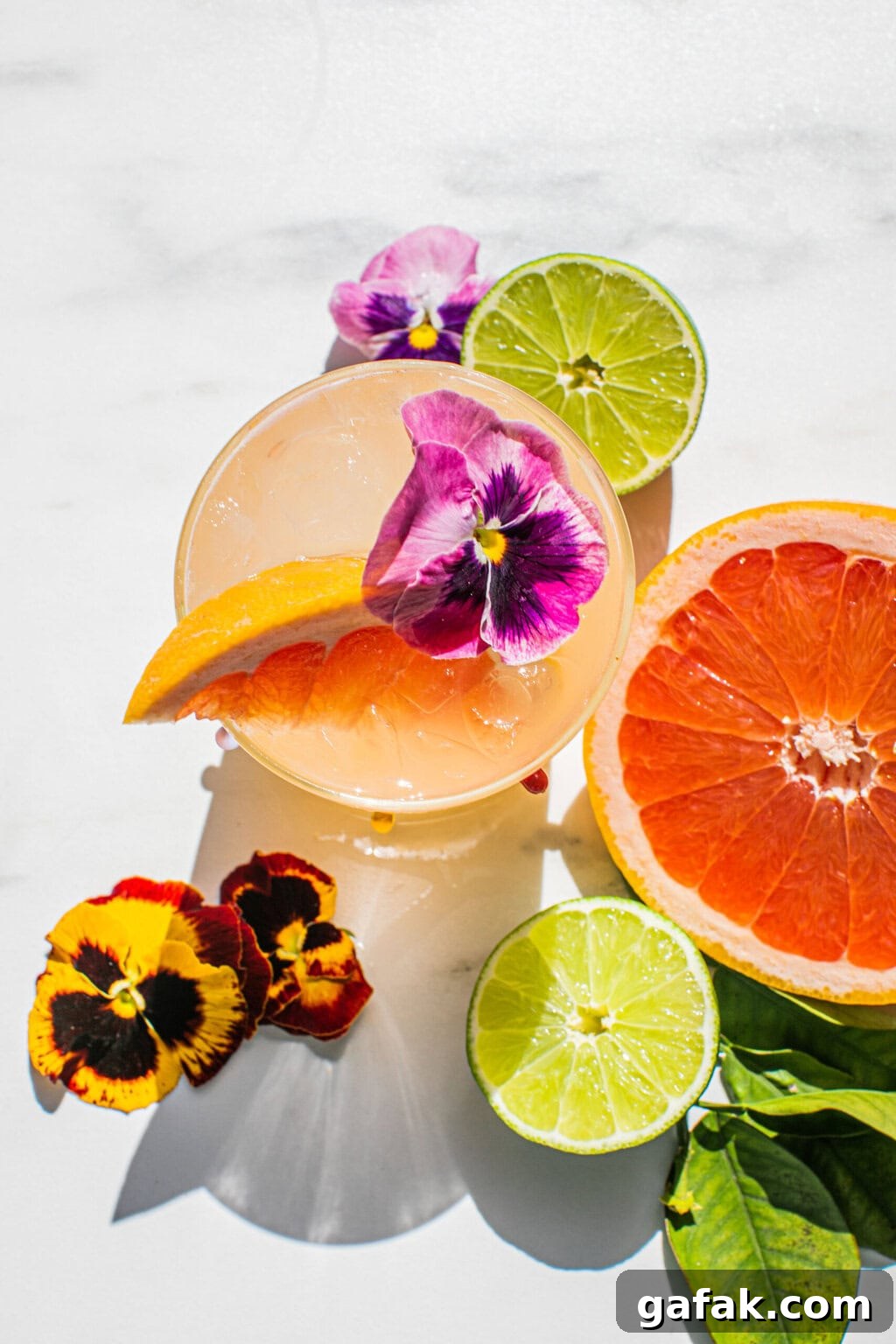 Make-Ahead Paloma Pitcher Cocktail Recipe 3 an overhead photo of a paloma garnished with edible flowers, a grapefruit wedge, and fresh lime and grapefruit scattered around it