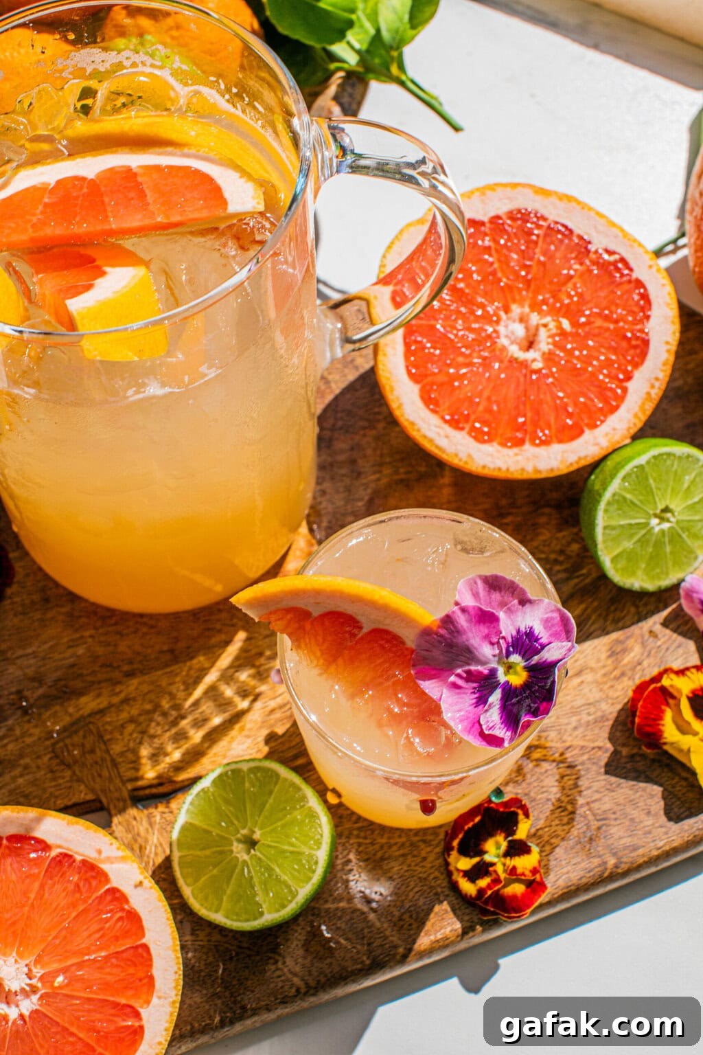Make-Ahead Paloma Pitcher Cocktail Recipe 8 a pitcher of palomas beside a glass of the drink garnished with edible flowers and a grapefruit wedge