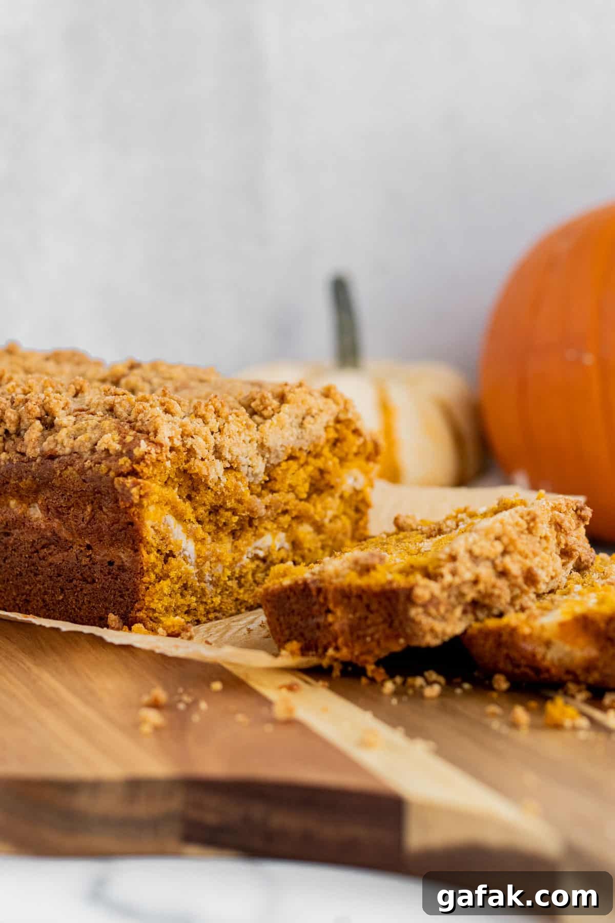 loaf of pumpkin cream cheese bread sitting on a wood cutting board with 2 slices cut. 2 pumpkins in the background.