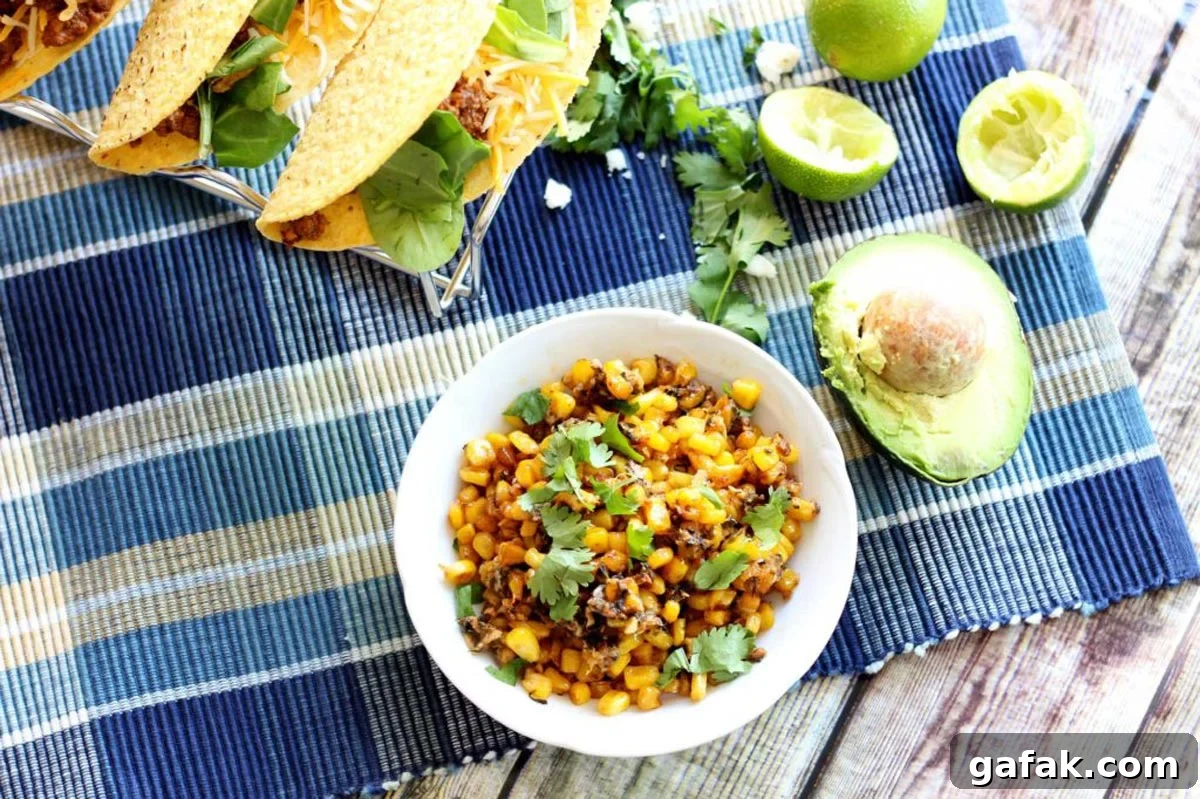 Authentic Mexican Street Corn (Elote) Recipe — Grilled & Creamy 8 White bowl containing Mexican Street Corn sitting on a blue tablecloth, tacos, cheese, avocado and limes on table.
