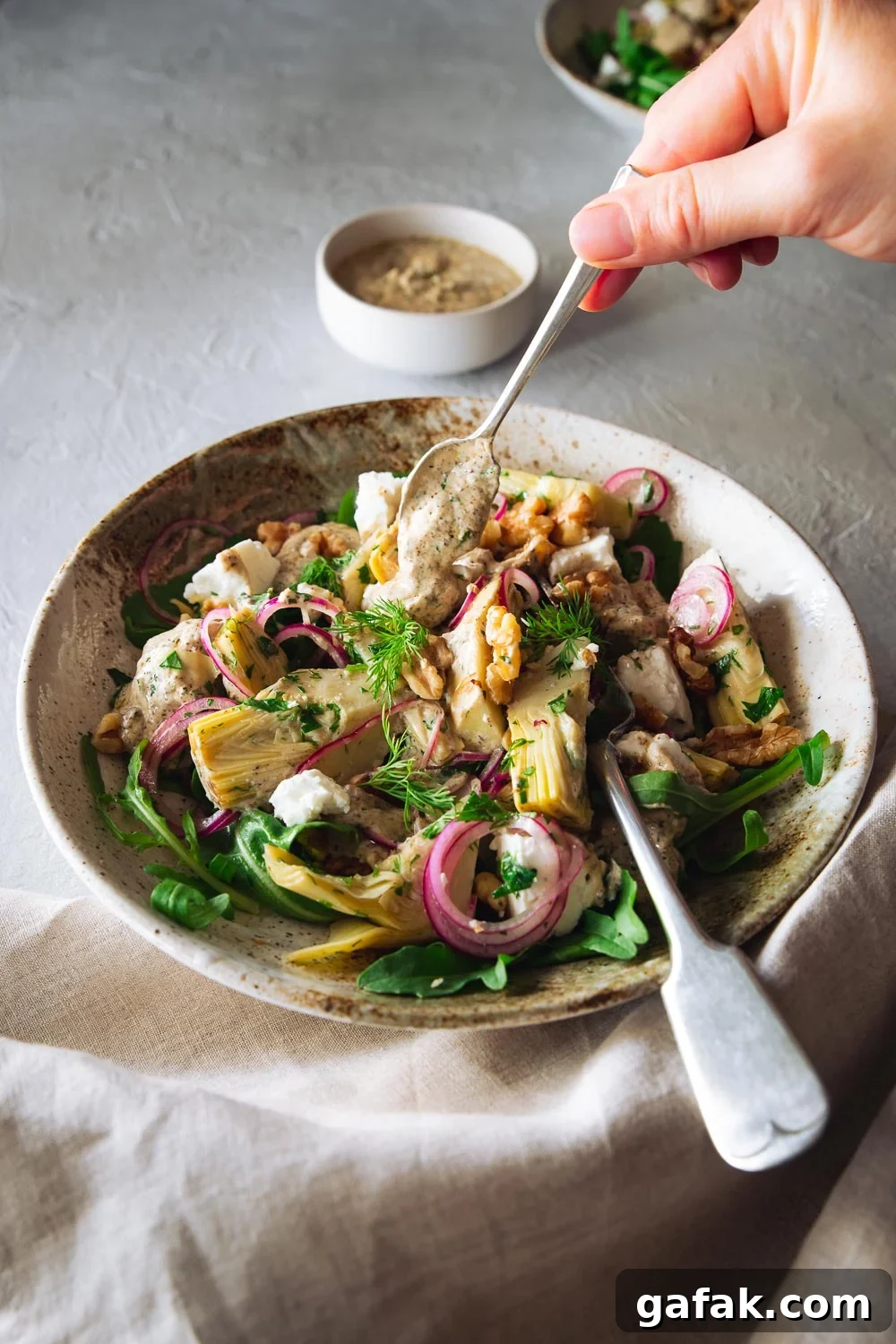 A hand drizzling yoghurt dressing over an artichoke salad in a salad bowl.