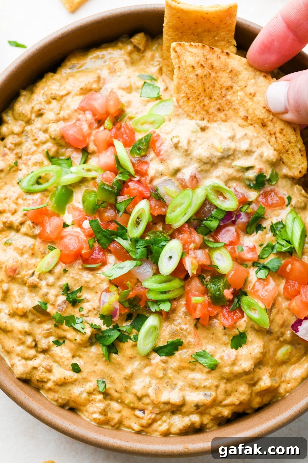 A large ceramic bowl filled with dairy free queso fundido with ground beef, topped with pico de gallo, cilantro, and thinly sliced green onions with a hand dipping a chip into the dip.