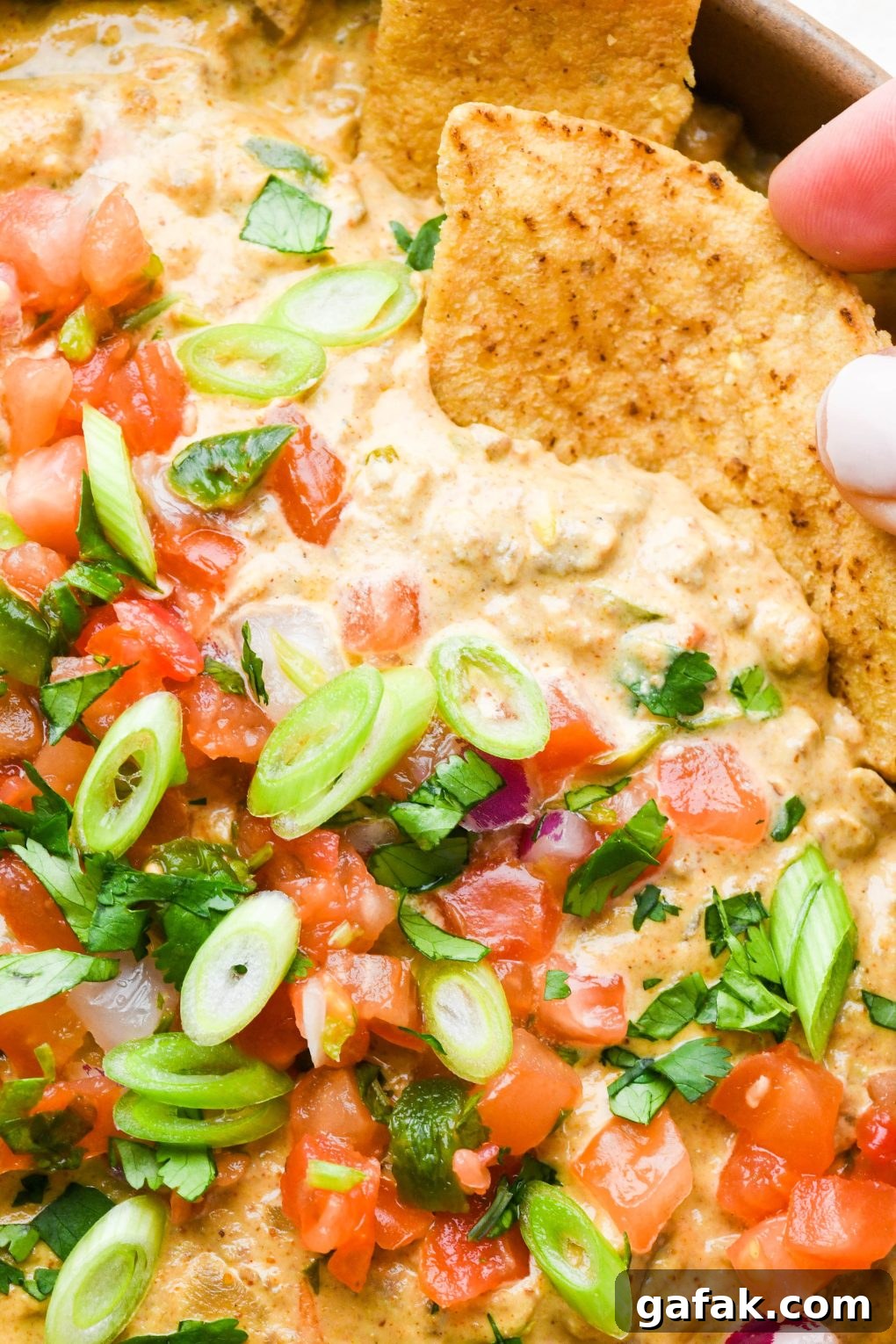 Close up of a large ceramic bowl filled with dairy free queso fundido with ground beef, topped with pico de gallo, cilantro, and thinly sliced green onions with a hand dipping a chip into the dip.