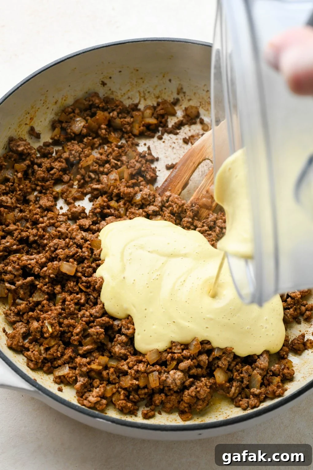 How to make Dairy Free Queso Fundido with Ground Beef: Cooked ground beef and onion in skillet with spices mixed in, pouring cashew queso into the skillet.