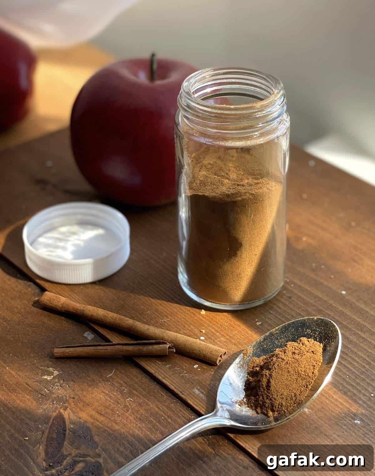 An open jar of apple pie spice mix with some on a spoon and cinnamon sticks.