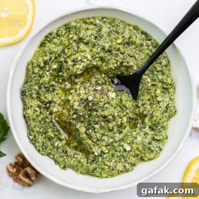 A bowl of vibrant green basil walnut pesto shown with lemon slices and garlic cloves next to the bowl.