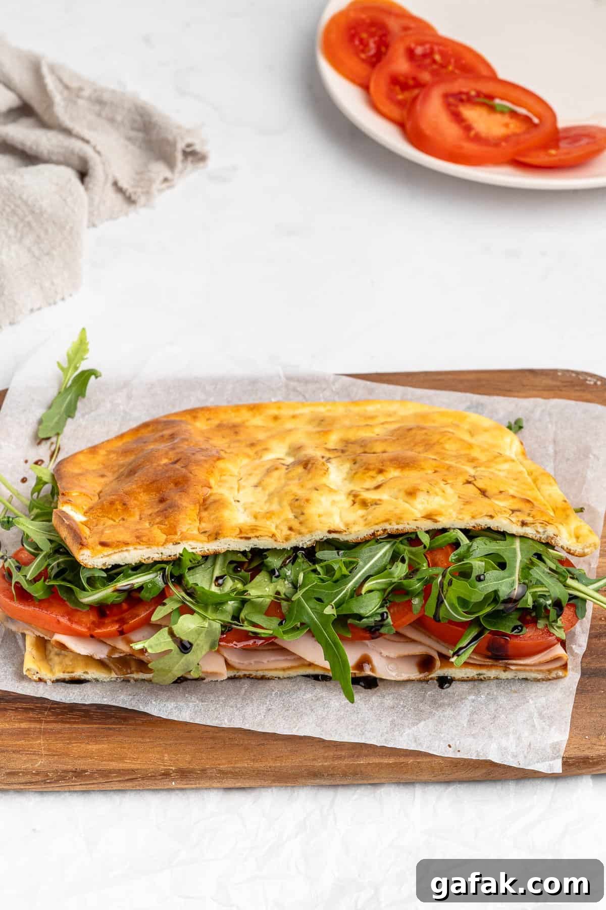 A sandwich made with focaccia bread, filled with turkey slices, fresh tomatoes, and arugula, sits on parchment paper on a wooden cutting board. A plate with sliced tomatoes is in the background, next to a beige cloth.