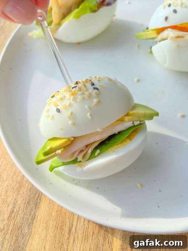 A hand holds a toothpick, securing an Egg Sandwich made with egg halves as buns, filled with turkey, cheese, lettuce, and avocado, garnished with sesame seeds—all served on a white plate.