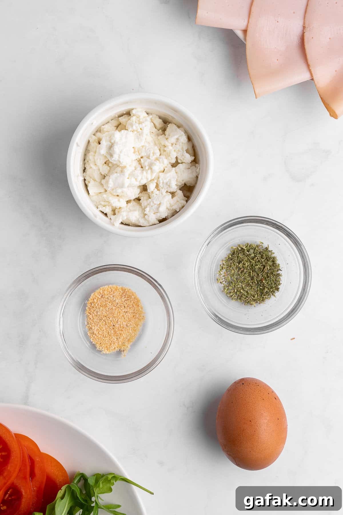 Top-down view of ingredients on a white surface: a bowl of ricotta cheese, dried herbs, garlic powder, sliced ham, a brown egg, fresh tomato slices, and arugula leaves.