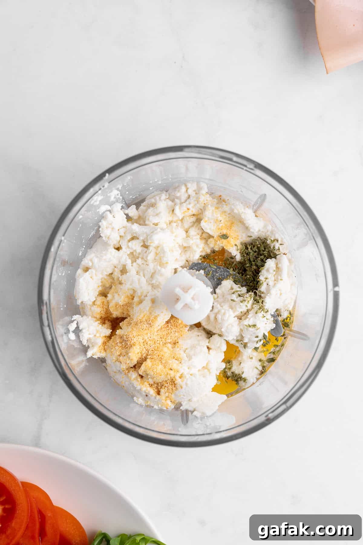 A food processor filled with ricotta cheese, breadcrumbs, herbs, and seasoning. Below, a plate with sliced tomatoes and basil is partially visible. The background is a white marble surface.