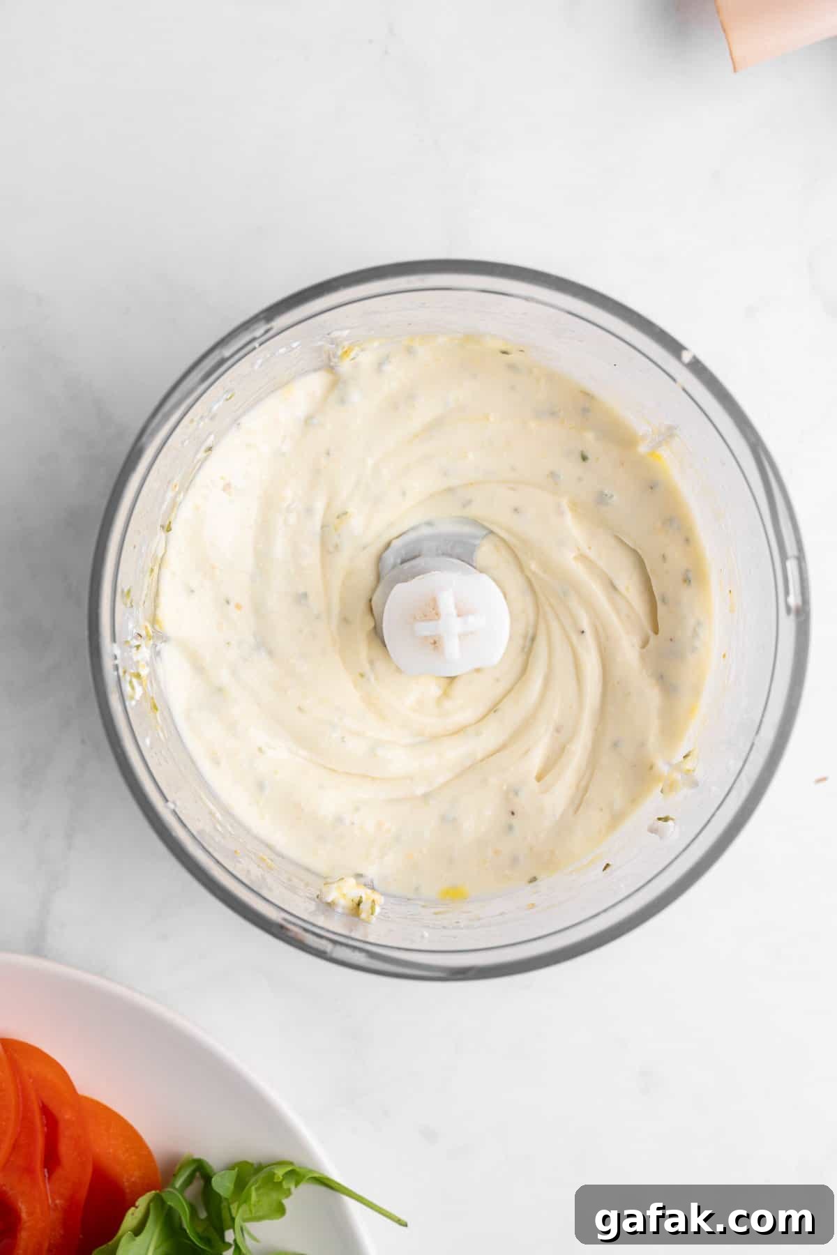 A food processor containing creamy, light-colored dressing with visible herbs. Next to it is a plate with sliced tomatoes, and a few leaves of fresh basil on a white marble surface.