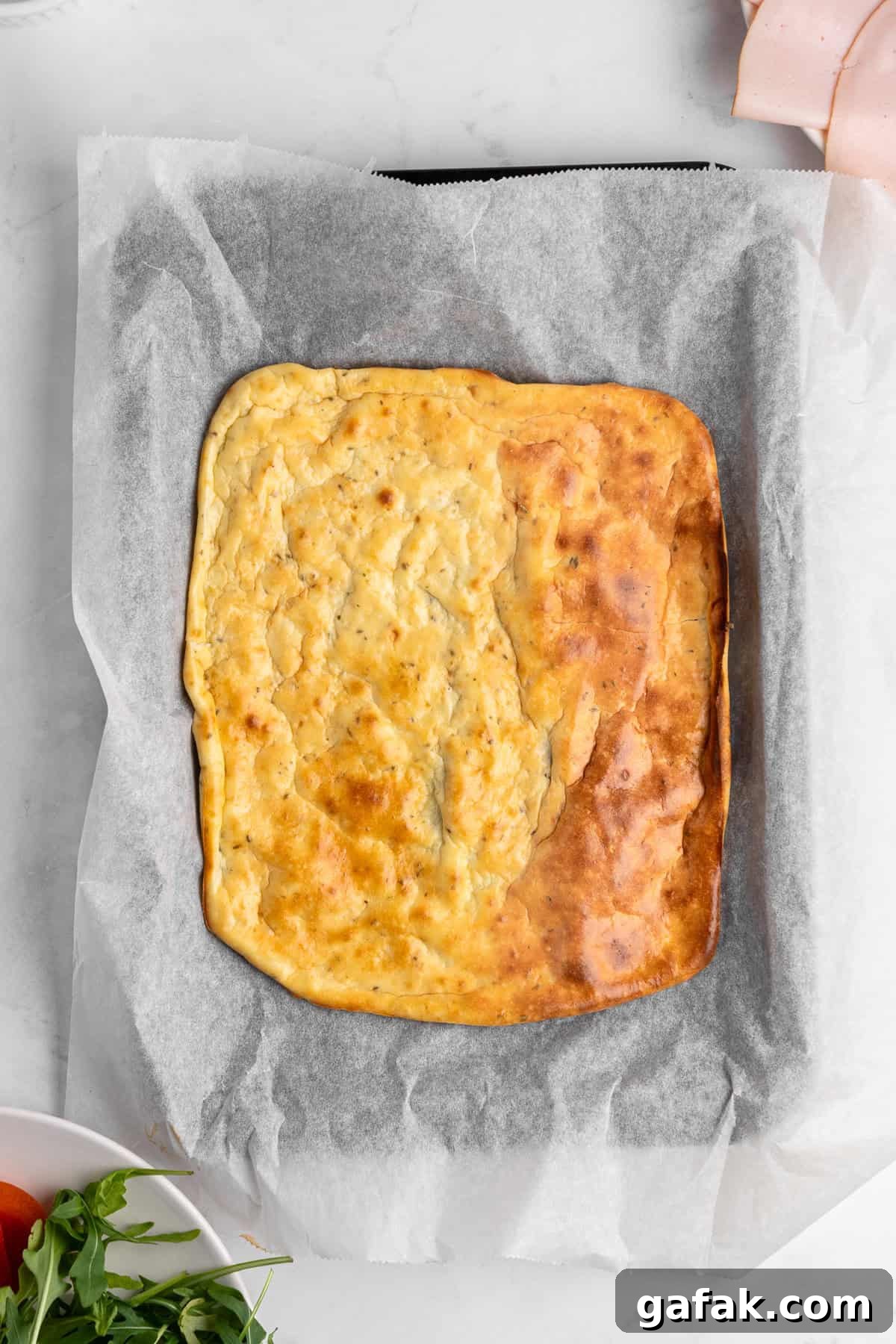 A freshly baked rectangular cottage cheese flatbread on parchment paper, resting on a marble surface. Ingredients like arugula, tomatoes, and slices of ham are partially visible around the quiche.