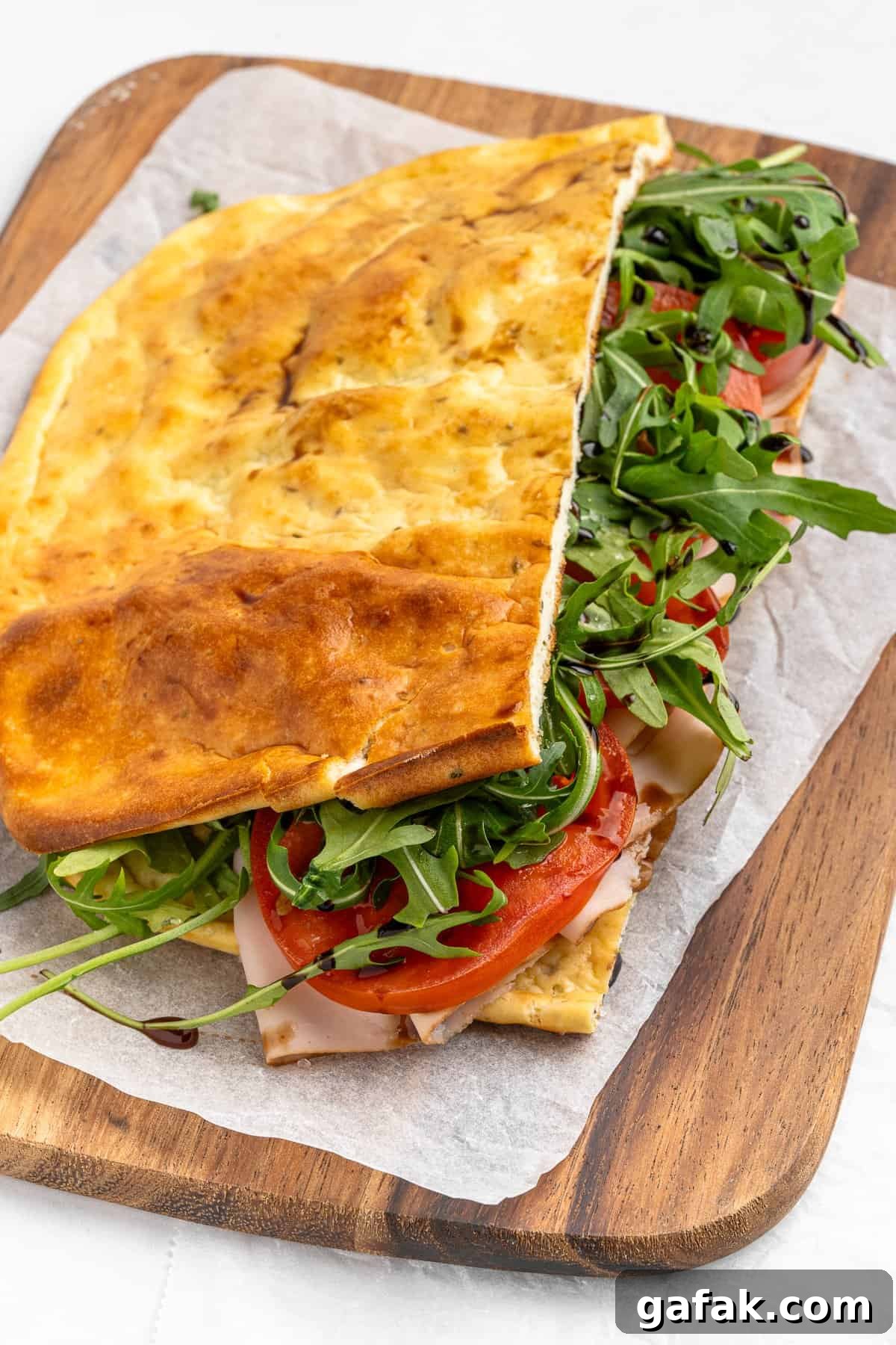 A folded flatbread sandwich on a wooden board, filled with sliced turkey, fresh tomato, arugula, and cheese. The bread appears golden and lightly toasted, resting on a piece of parchment paper.