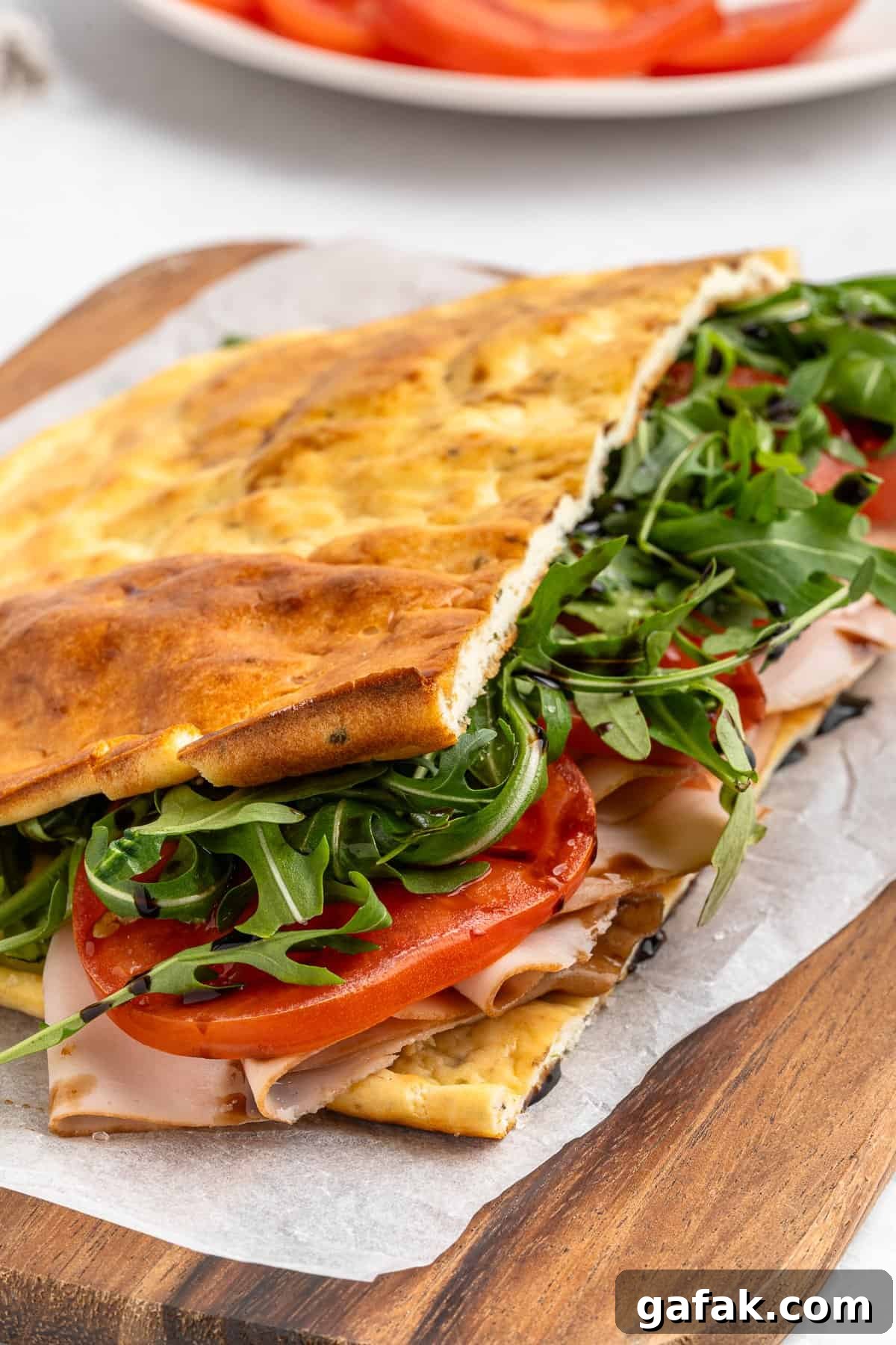 A sandwich on a wooden board, made with flatbread, filled with arugula, sliced tomatoes, and deli turkey. The flatbread is folded over the ingredients, which are visible. A plate with more tomato slices is in the background.