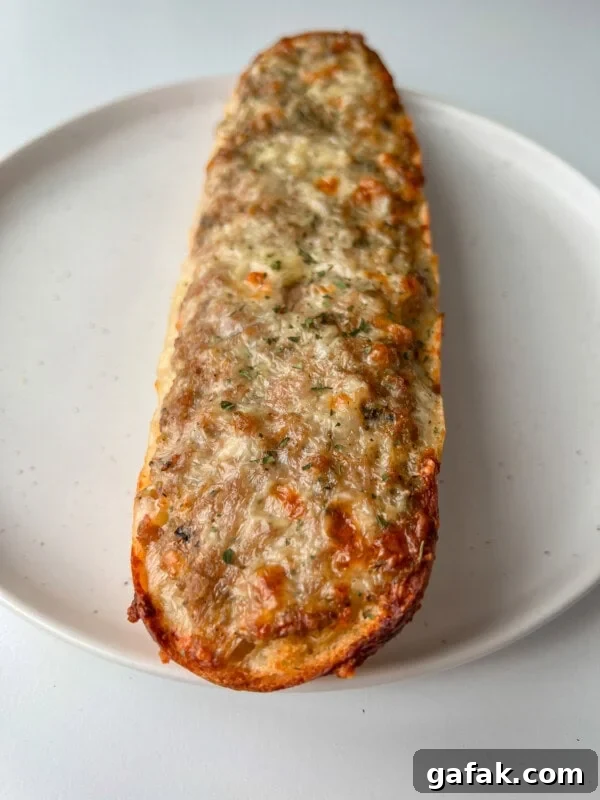 A close-up of a baked, cheese-covered piece of garlic bread on a white plate, with melted, slightly browned cheese and herbs sprinkled on top.