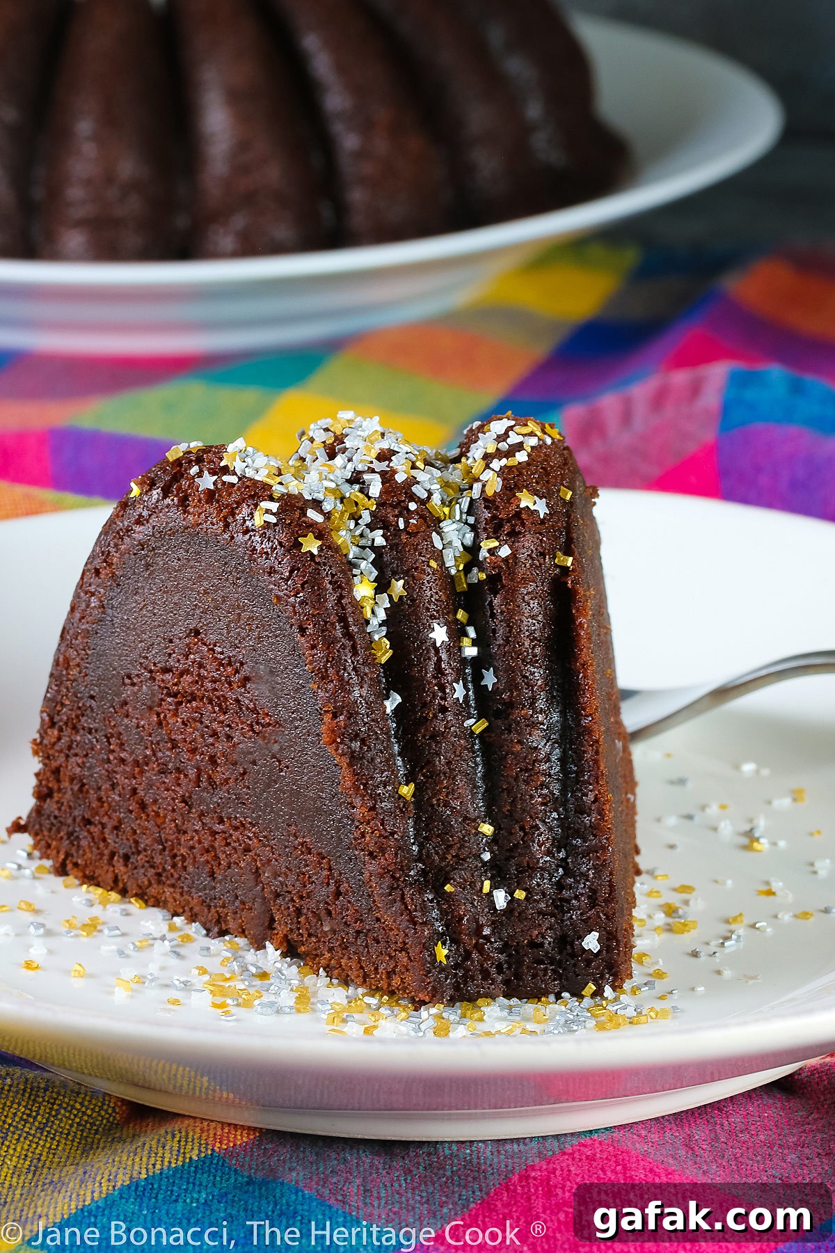 Slice of chocolate bundt cake on white plate in front of the remaining whole cake, topped with silver and gold sprinkles like a snow shower