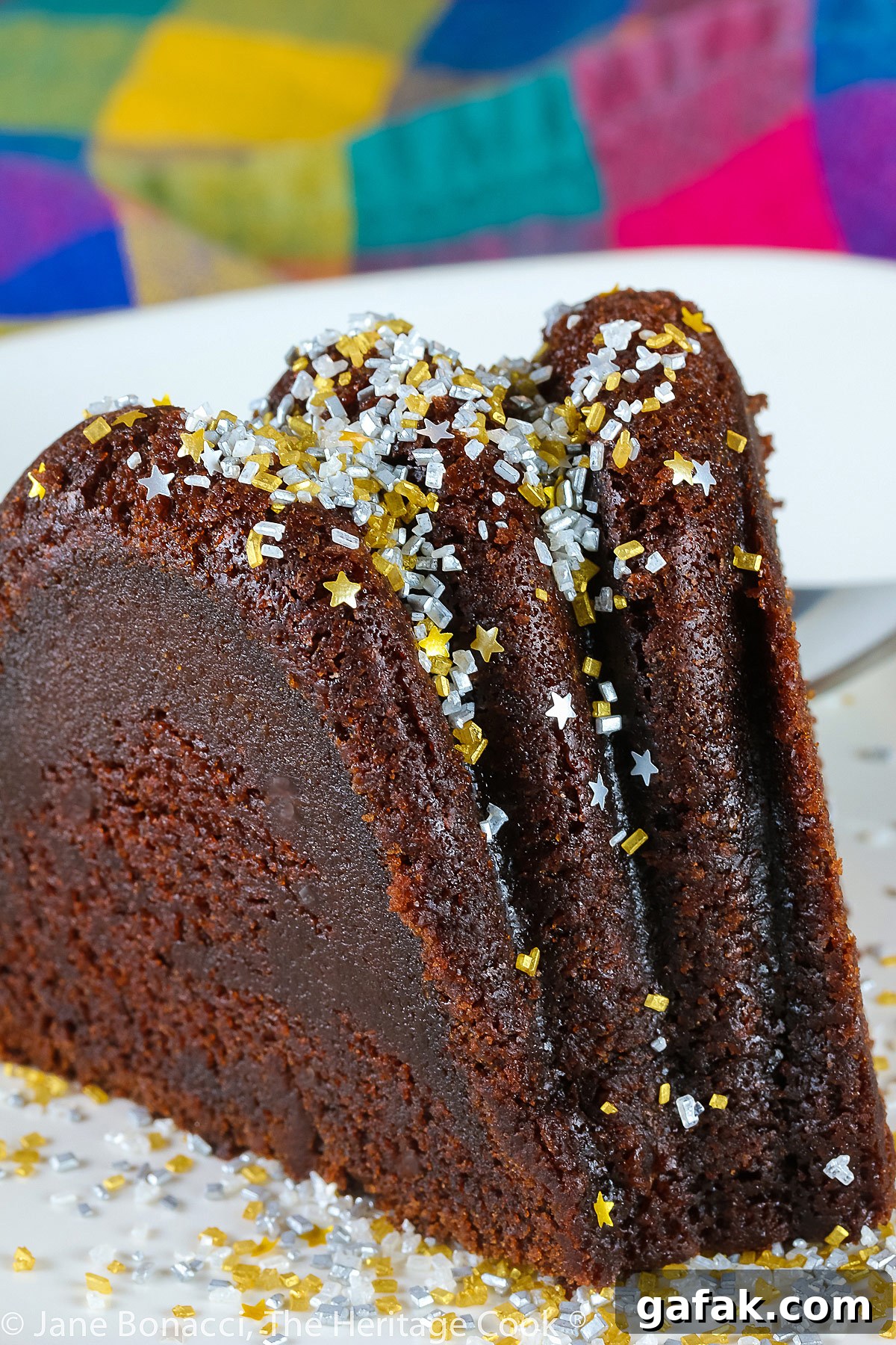 Slice of chocolate bundt cake on white plate in front of the remaining whole cake, topped with silver and gold sprinkles