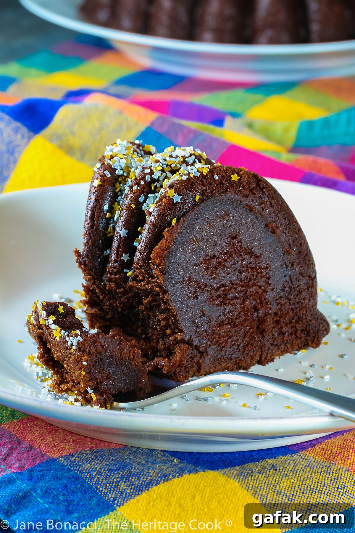 Slice of chocolate bundt cake on white plate in front of the remaining whole cake, topped with silver and gold sprinkles