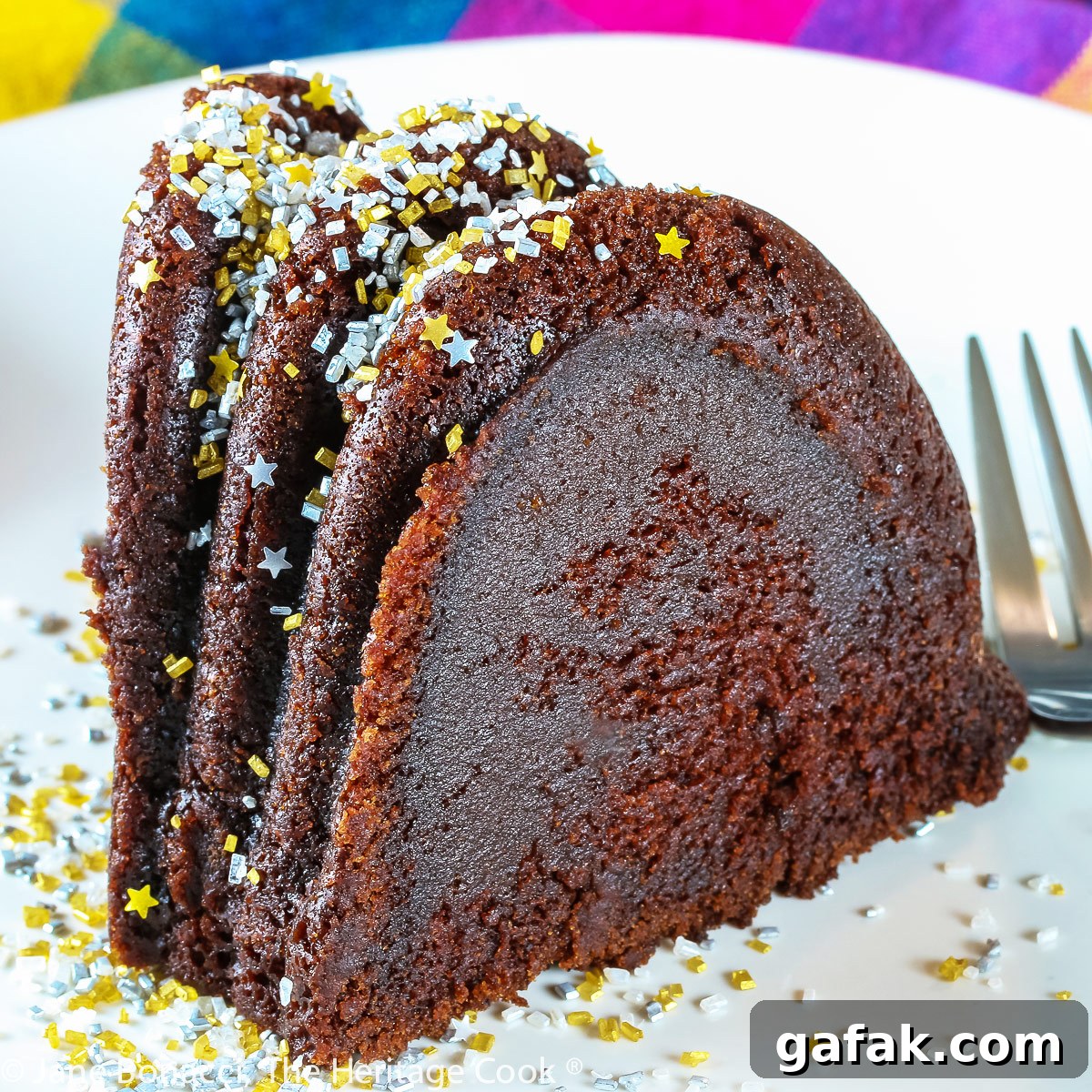 Slice of chocolate bundt cake on white plate in front of the remaining whole cake, topped with silver and gold sprinkles