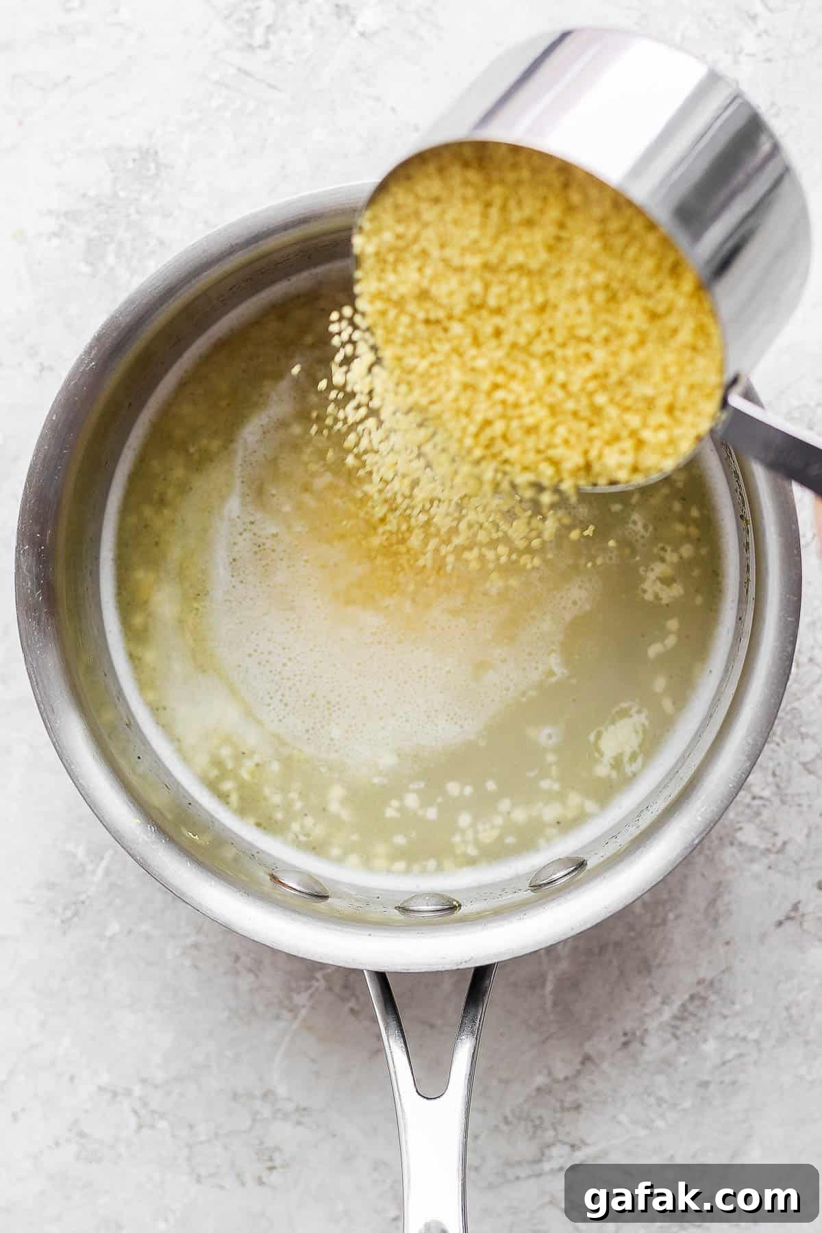 A cup of raw couscous being poured into the water mixture in the sauce pan.