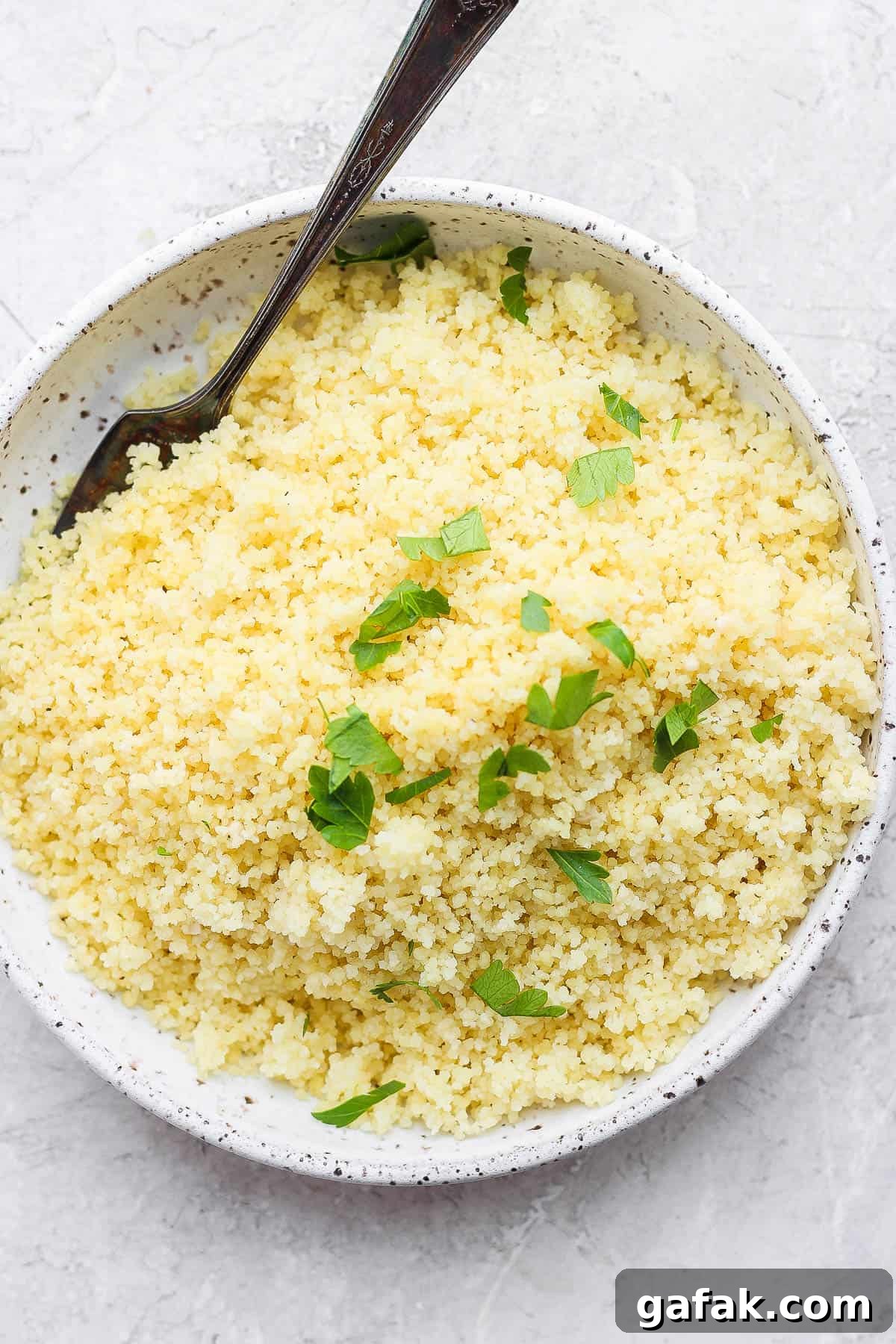Cooked couscous in a serving bowl and sprinkled with chopped parsley.  A fork is off to the side of the bowl.