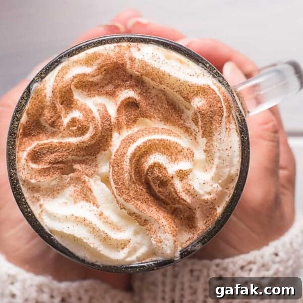 A person holding a glass mug of whipped cream-topped cinnamon latte, sprinkled with cinnamon.