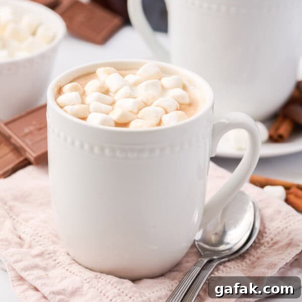 A white mug filled with hot chocolate topped with mini marshmallows, placed on a pink cloth with a spoon beside it.