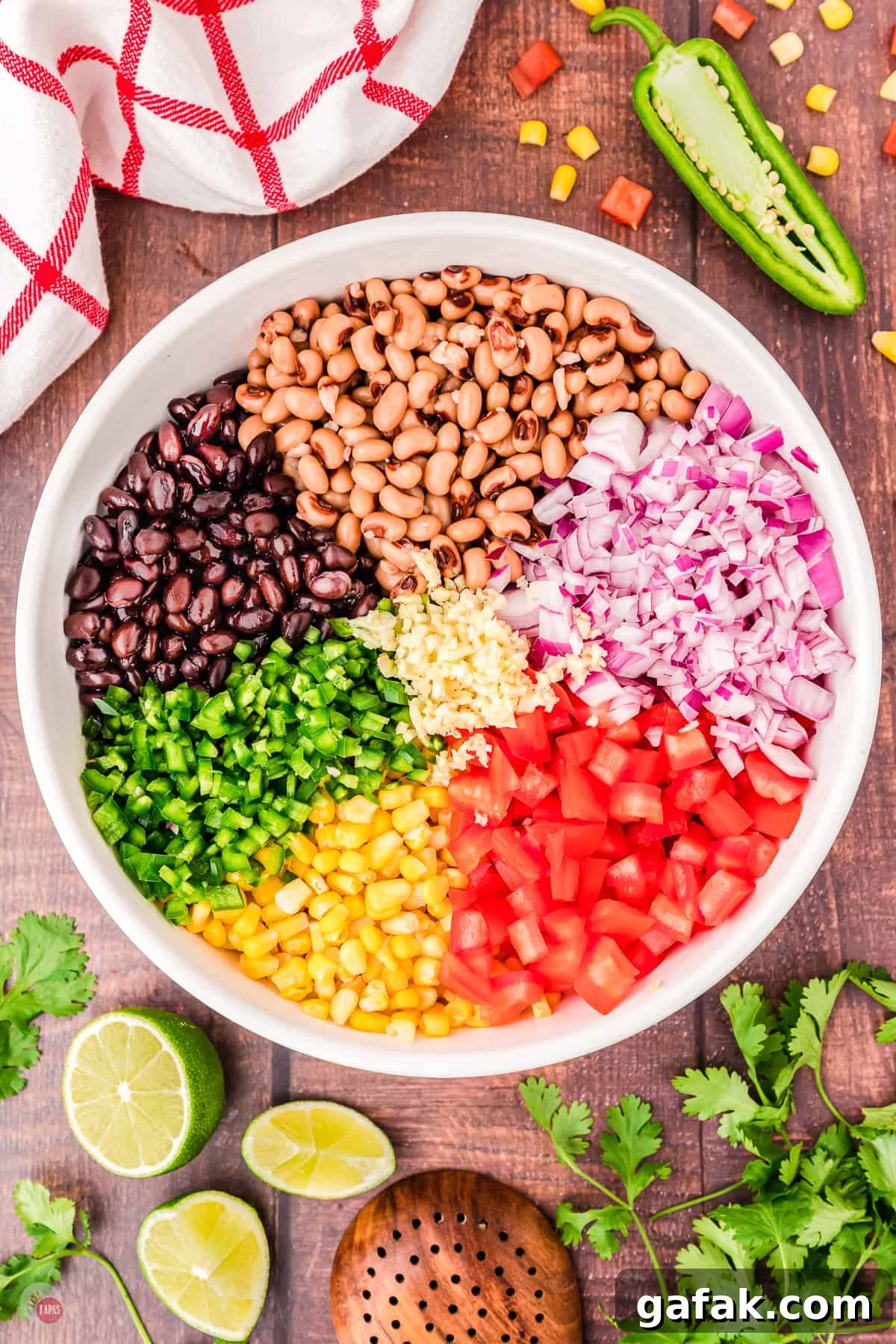 Top view of large white bowl on a table with all the chopped ingredients of cowboy caviar in it in sections, with some limes, cilantro and a jalapeno on the table next to it. 