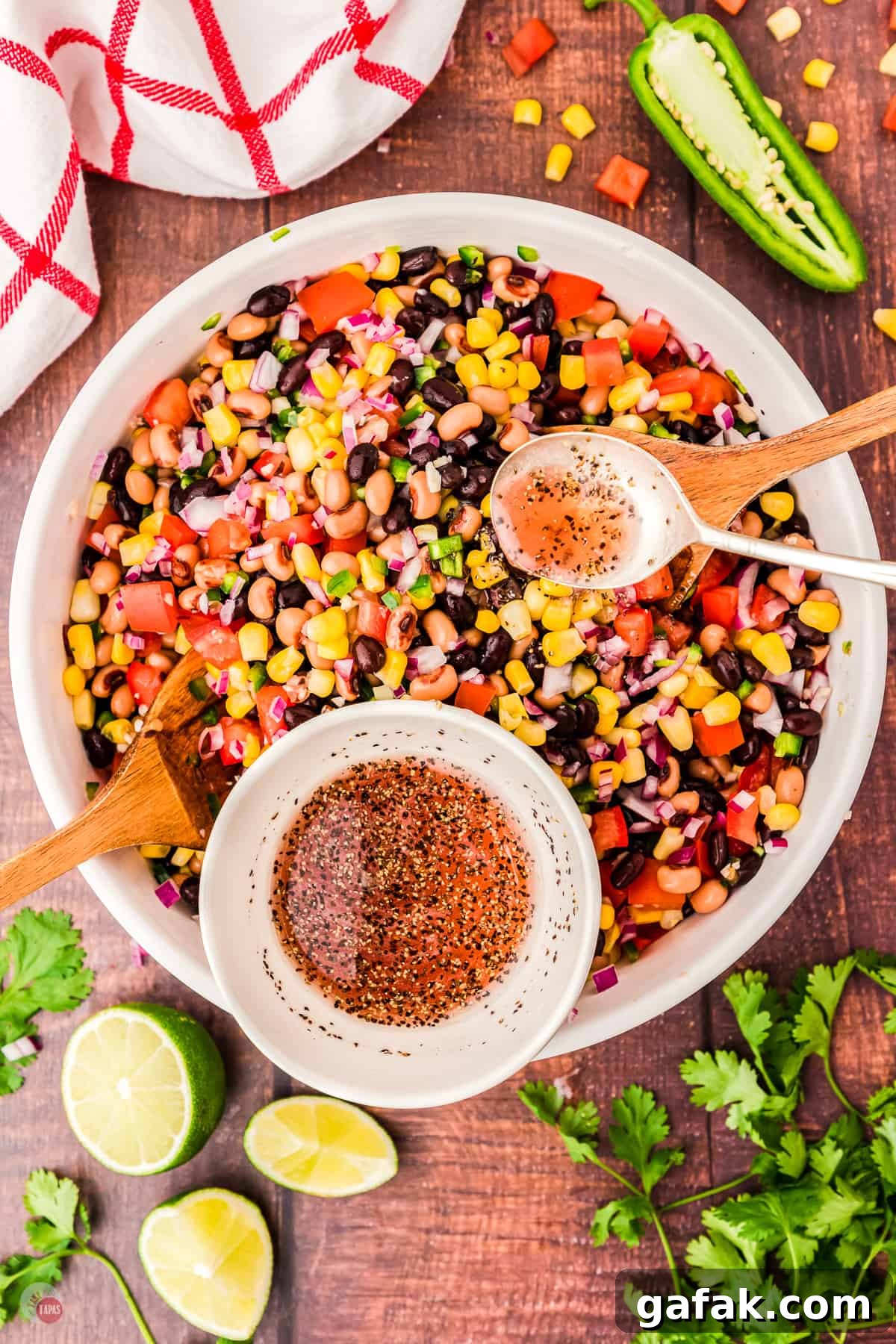 Top view of large white bowl on a table with all the chopped ingredients of cowboy caviar mixed up, with a white ramekin sitting on top of it with the dressing in it. There are also some limes, cilantro and a jalapeno on the table next to the bowl. 