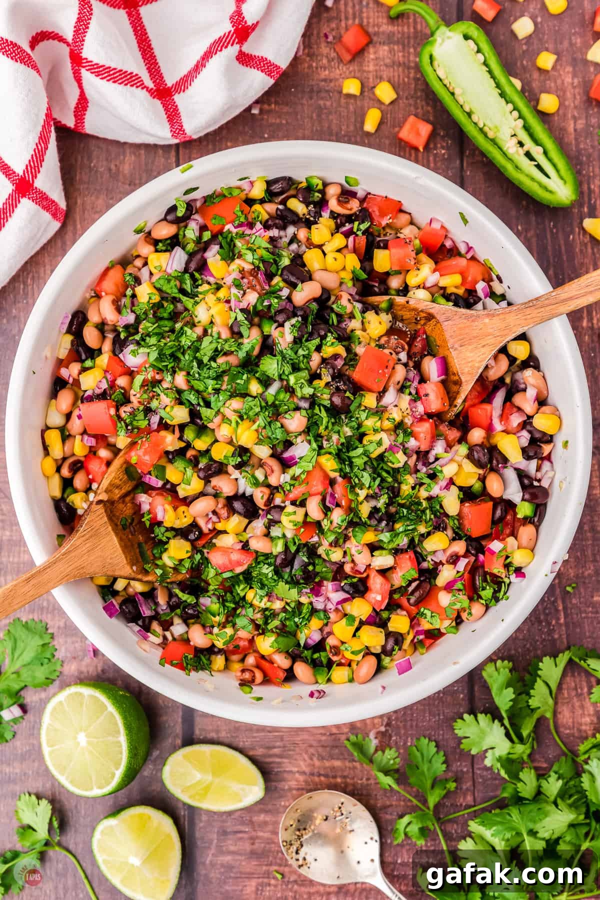 Top view of a large white bowl filled with cowboy caviar salad with two wooden serving utensils in it on either side. There are also some limes, cilantro and a jalapeno on the table next to the bowl. 