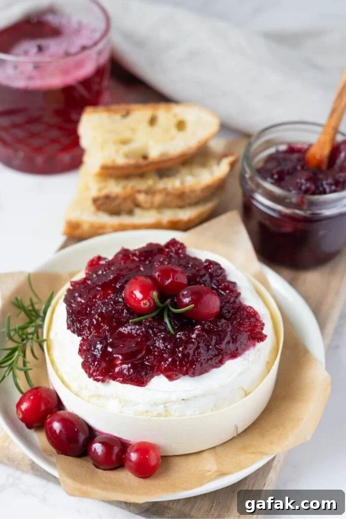 A wheel of Brie topped with vibrant cranberry vanilla jam. The Brie is decorated with fresh cranberries and rosemary and you can see the jar of jam and stack of sourdough bread slices in the background.