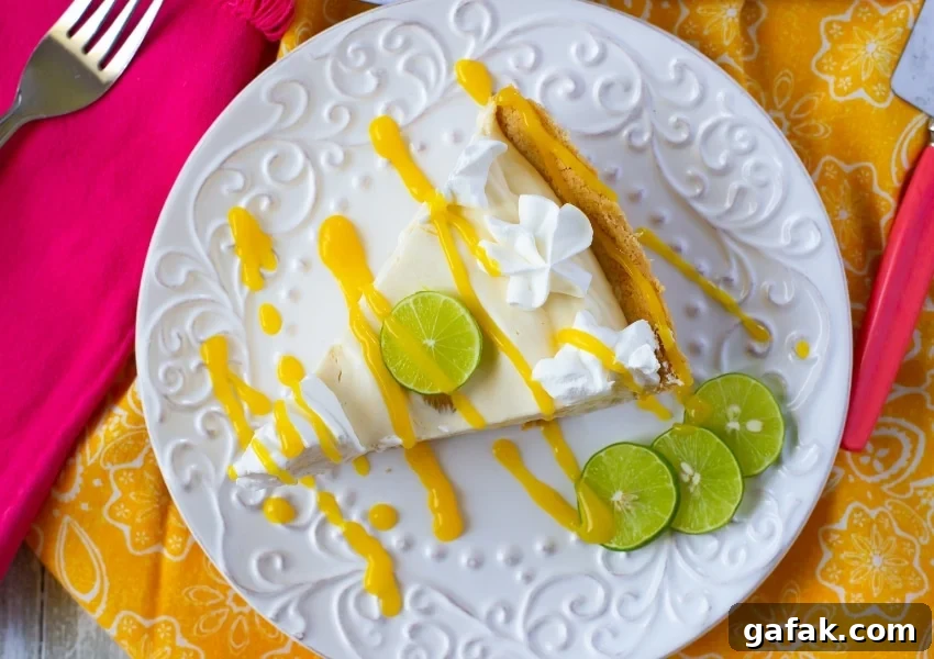 Above the plate photo of a slice of Mango Key Lime Pie that has been garnished with whipped cream, a slice of lime and a drizzle of mango sauce on top of a yellow cloth napkin.
