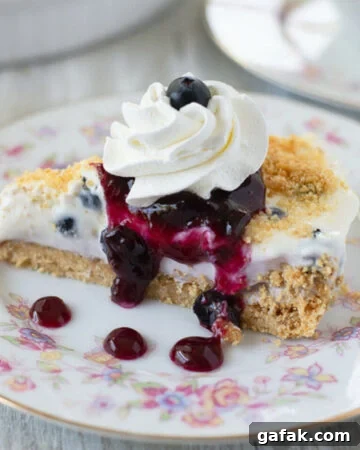 sliced no bake blueberry cheesecake pie on a floral patterned dessert dish with whipped cream and blueberry sauce drizzled over it.