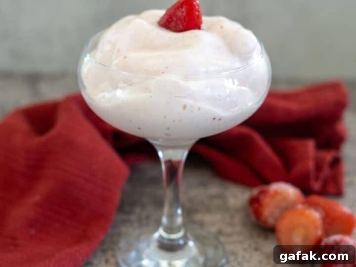 A dessert glass filled with strawberry mousse garnished with a strawberry slice, placed on a textured surface beside a red cloth and some whole strawberries.