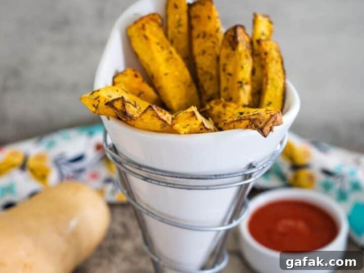A white cone-shaped container holds butternut squash fries seasoned to perfection. A small bowl of tantalizing dipping sauce awaits in the background.
