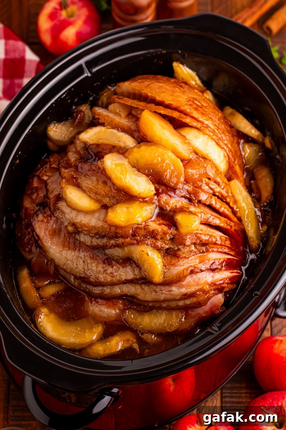 looking down at wooden table with a black slow cooker with apple glazed spiral ham. Fresh apples around table 